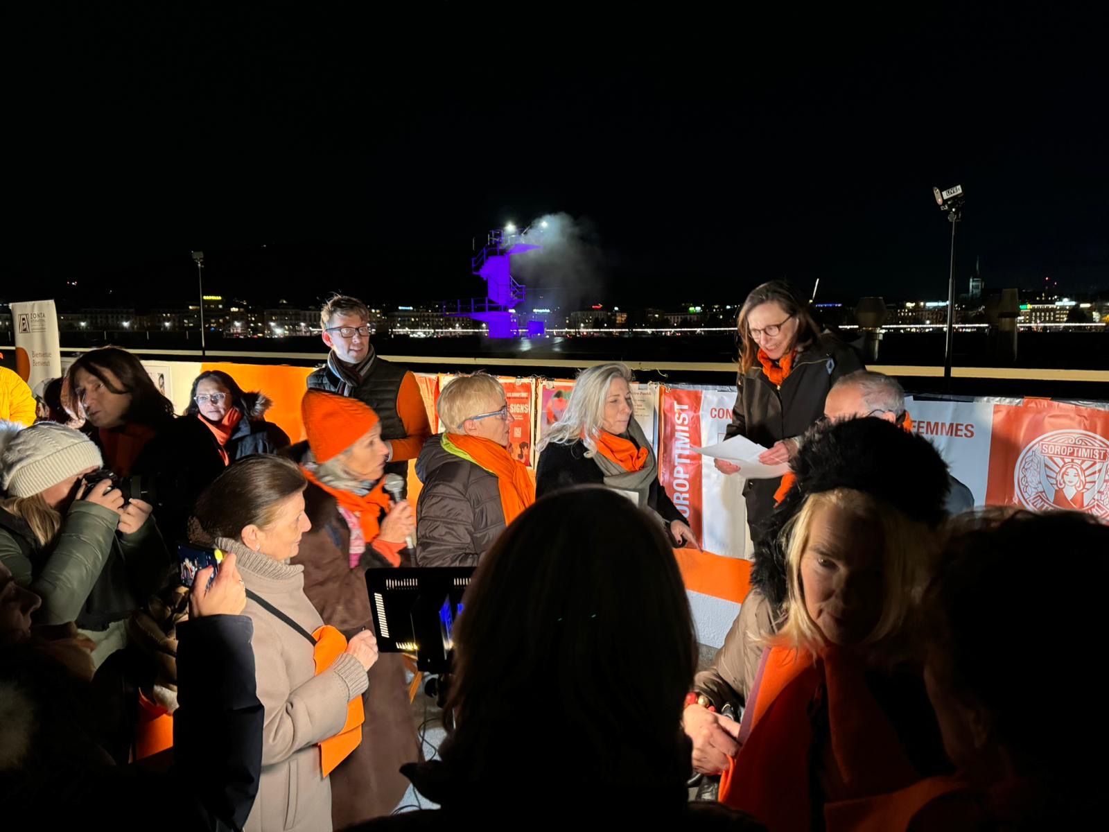 Speakers in orange scarves addressing the crowd at night by the lake in Geneva